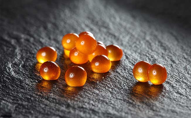 "Close-up macro shot of glistening, individual bright orange Priess & Co. Danish trout caviar pearls scattered on a dark, textured slate surface, highlighting their translucent quality."