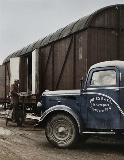 "Historical photo of a dark blue Priess & Co. fish export truck, marked 'PRIESS & Co. Fiskeexport Glyngøre,' parked next to a large wooden train wagon. A worker stands on a ramp, actively loading or unloading goods, illustrating the company's early operations in Glyngøre." width="430" height="554"