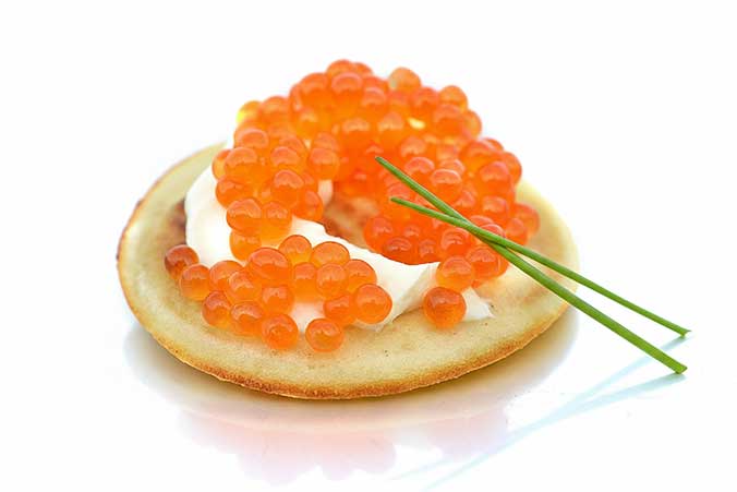 "Close-up of golden potato blini topped generously with vibrant orange Priess & Co. trout caviar and fresh thyme sprigs, served as an elegant appetizer." width="676" height="451"