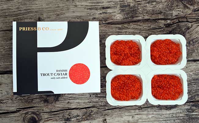 "Overhead view of Priess & Co. Danish trout caviar SmartPack: a white box with the brand logo, alongside the convenient 4x50-gram portion packaging filled with vibrant orange caviar, on a rustic wooden background."